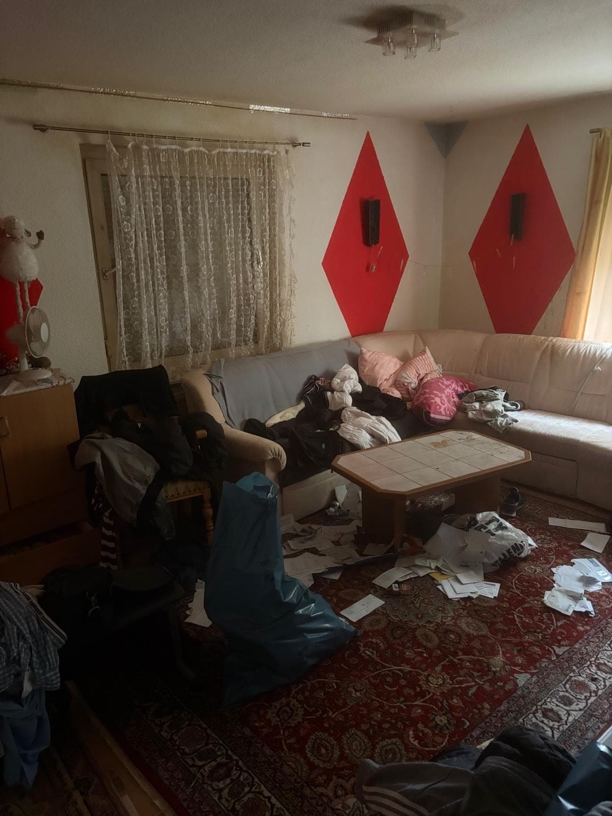 Wohnzimmer vor der Räumung: Ecksofa mit Kleidung, dazu Tisch, Papiere und ein Müllsack auf dem Teppich